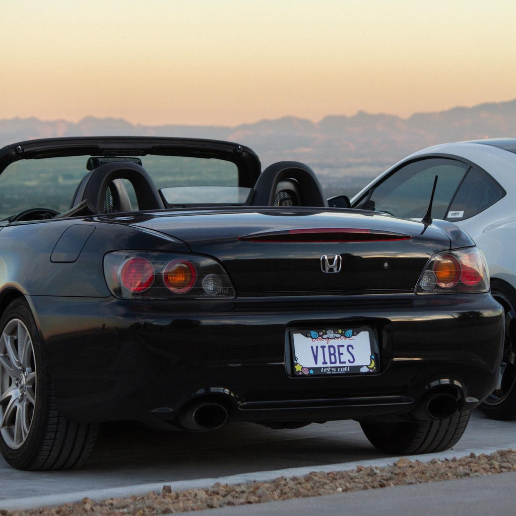 Black Honda S2000 with 'VIBES' license plate on a scenic road at sunset.